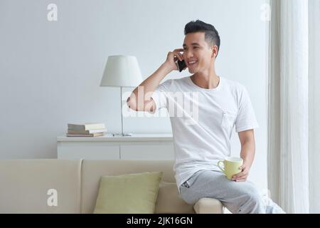 Hübscher junger asiatischer Mann mit einer Tasse Tee, der auf dem Sofa sitzt und am Telefon anruft Stockfoto