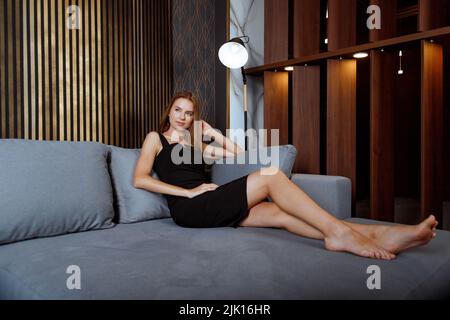 Junge Frau in schwarzem formellen Kleid entspannen Sie sich auf dem bequemen Sofa zu Hause entlasten negative Emotionen Stockfoto