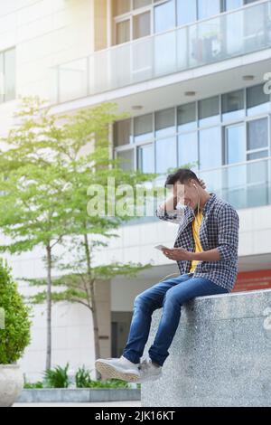 Glücklicher asiatischer junger Mann, der Musik über Anwendung in seinem Telefon hört Stockfoto
