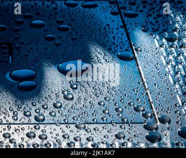 Große bis kleine Wasserperlen auf einer strukturierten Metalloberfläche, die von hellblau auf dunkelblauen Hintergrund abfärbt. Stockfoto