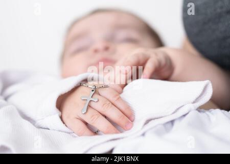 Nahaufnahme der Hand eines Babys bei seiner Taufe mit einem christlichen Kreuz. Junge Mutter hält ihr Baby bei ihrer katholischen Taufe in der lateinischen Kirche. cau Stockfoto