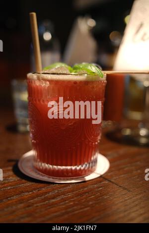 Glas gefüllt mit rotem Cocktail auf einem Holztisch mit verschwommenem Licht im Hintergrund Stockfoto