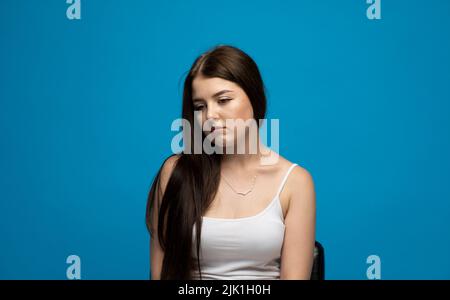 Portrait von verärgert junge beautifil Brünette Mädchen mit langen Haaren in einem weißen T-Shirt stehen mit geschwollenen Wangen isoliert auf blauem Hintergrund. Emotion von Stockfoto