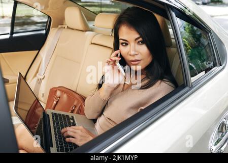 Wunderschöne junge Frau mit Laptop im Auto und sprechen über das Telefon Stockfoto