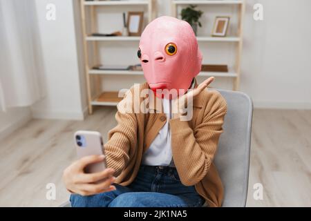 Glücklich lustige blonde Dame in rosa Fischmaske warmen Pullover tun Selfie Video-Anruf sitzen im Sessel im modernen Zuhause Interieur. Pause von der Arbeit, nehmen Sie ein Stockfoto