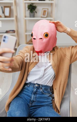 Glücklich lustige blonde Dame in rosa Fischmaske warmen Pullover tun Selfie Video-Anruf sitzen im Sessel im modernen Zuhause Interieur. Pause von der Arbeit, nehmen Sie ein Stockfoto