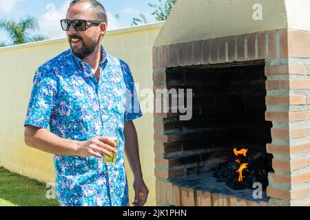 Junger Hipster-Mann mit Bart, Sonnenbrille und hawaiiihemd trinkt ein Bier, während er ein Barbecue für das Essen im Ferienchalet im Sommerurlaub vorbereitet Stockfoto