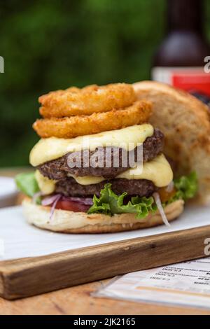 Doppelter Cheeseburger mit Zwiebeln, Tomatensalat, Cheddar und Zwiebelringen auf einem Holzbrett Stockfoto