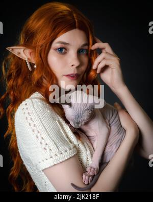 Junge weibliche Elfe cosplay mit schlafenden Sphinx Kätzchen. Nahaufnahme Porträt auf schwarzem Hintergrund. Niedliche Elfe in weißem Strickkleid, mit langen roten Lockenhaaren Stockfoto