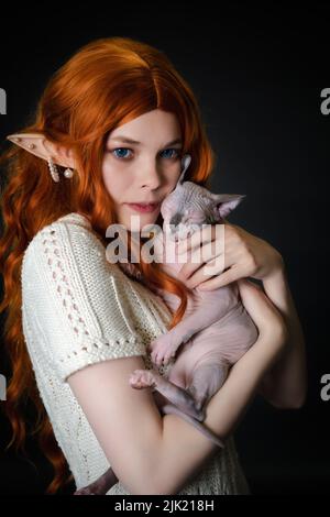 Porträt der Elfen Cosplay junge Frau in weißem Strickkleid sorgfältig umarmt schlafenden Sphinx Pussycat. Schwarzer Hintergrund. Rotschopf-Elf schaut weg Stockfoto