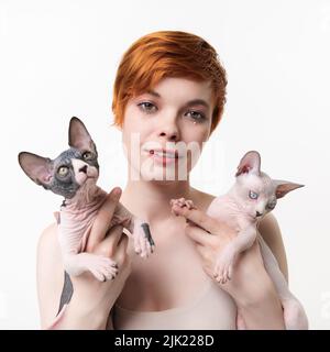 Rotschopf junge Frau umarmt zwei Sphynx Katze in den Händen auf weißem Hintergrund. Hübsche Frau mit kurzen Haaren und Blick auf die Kamera. Quadratisches Format, Teil einer Serie Stockfoto