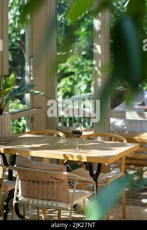 Weinkelch auf dem Esstisch. Weiches Licht mit Schatten. Großes Fenster mit Blick auf die umliegende Natur. Stockfoto