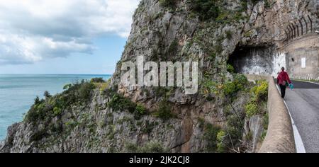 Eine weibliche Tourist zu Fuß auf der Straße an der Amalfiküste, die alte historische Weise ist auf der linken Seite der Straße, Italien überwuchert gesehen Stockfoto