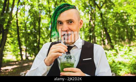 Junger Mann mit Dreadlocks trinkt erfrischende Limonade im Waldpark. Lustiger Hipster mit grüner Frisur trinkt Softdrink durch Stroh, abschreckend Stockfoto