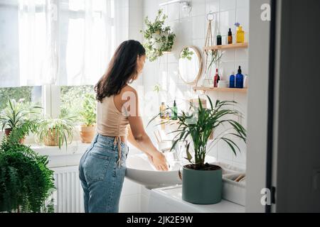 Junge schöne Frau mit dunkler Haut waschen Hände in hellem Bad mit vielen grünen Pflanzen. Biophile Gestaltung des Innenraums. Naturkosmetik, Selbstpflege und Wellness-Konzept Stockfoto