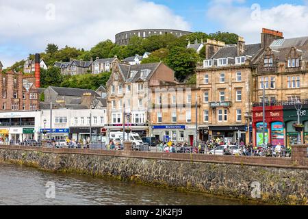 Oban Stadtzentrum an sonnigen Sommertagen, Geschäfte und Touristen in der Stadt mit Blick auf McCaig's Turm, Oban, Argyll, Schottland, Großbritannien Stockfoto