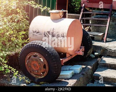 An einem sonnigen Sommertag steht ein hellrosa rostiger Eisentank auf Rädern auf Betonplatten auf dem Land. Wasserbehälter. Stockfoto