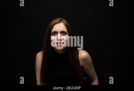 Portrait von verärgert junge beautifil Brünette Mädchen mit langen Haaren in einem schwarzen T-Shirt in einem schwarzen Raum. Emotion von wütend und aufgeregt. Stockfoto