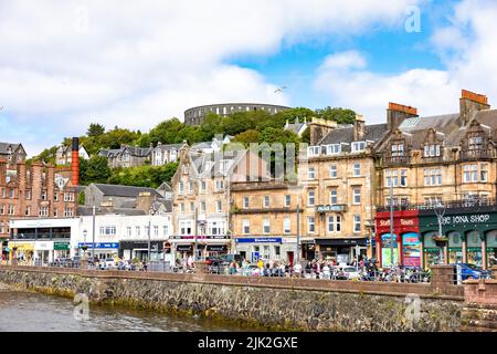 Oban Stadtzentrum an sonnigen Sommertagen, Geschäfte und Touristen in der Stadt mit Blick auf McCaig's Turm, Oban, Argyll, Schottland, Großbritannien Stockfoto