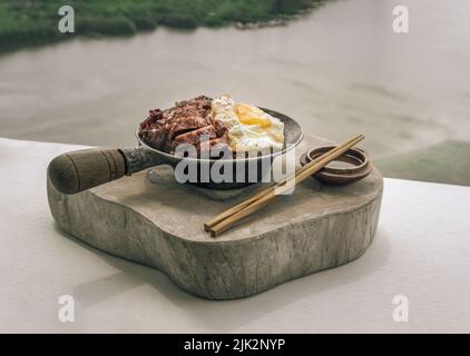 Reis mit rotem Grillfleisch, Spiegelei und gedünstetem pak-Choi-Kohl mit roter Sauce und Holzstäbchen auf Holzschneidebrett servieren. Asiatischer Essensstil Stockfoto