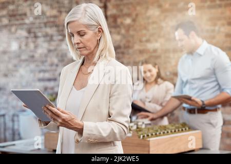 Reife Architektin, die ein Tablet verwendet, um Entwürfe in einem modernen Büro zu analysieren. Ältere Geschäftsfrau mit einem verschwommenen Hintergrund einer jungen Architektur Stockfoto