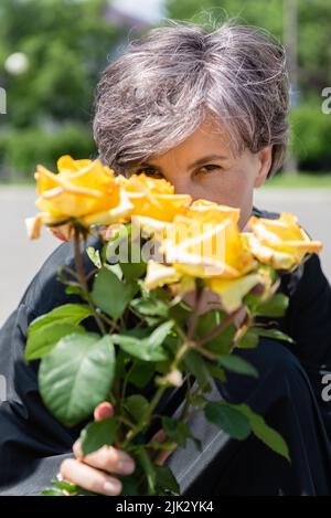Ein Strauß blühender, gelber Rosen von hinten, von der eine Frau mit Interesse in ihren Augen auf die Kamera schaut. Stockfoto