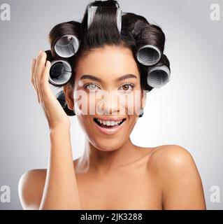 Ich bin begeistert von diesen Locken. Studioaufnahme einer attraktiven jungen Frau, die mit Lockenwicklern im Haar vor grauem Hintergrund posiert. Stockfoto