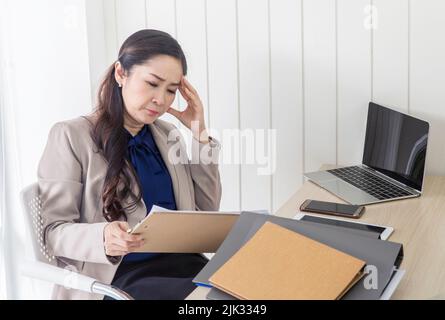 Asiatische Geschäftsfrau mit Kopfschmerzen im Büro, müde Buchhalterin mit Papiermappen im Büro, sich bei der Arbeit krank fühlend Stockfoto