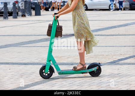 Eine junge Frau in einem leichten Kleid fährt mit einem gemieteten Elektroroller durch die Stadt. Speicherplatz kopieren. Das Konzept der umweltfreundlichen Bewegungsfreiheit. Stockfoto