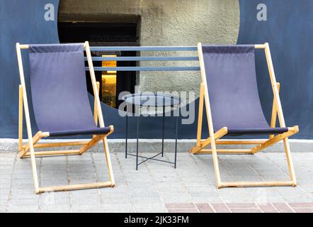 Zwei blaue Textil-Klappstühle mit einem Holzrahmen um einen Metalltisch vor dem Hintergrund eines gepflasterten Bürgerwegs und eines Café im Freien. Speicherplatz kopieren. Stockfoto