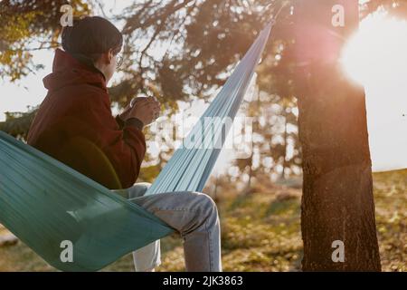 Hinten auf Frau mit kurzen Haaren, die Thermoskannen von Kaffee hält und in der Hängematte in Holz ruht. Stockfoto