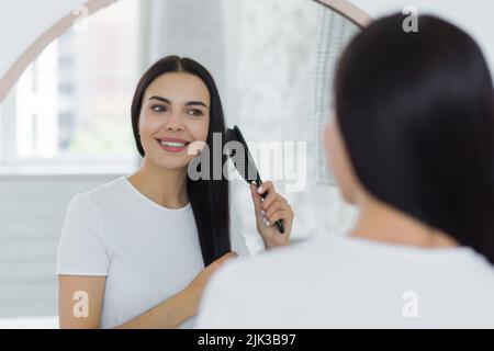 Schöne junge Frau, die morgens ihre Haare kämmte, in einen großen runden Spiegel schaute und lächelte, brünett in einem hellen Schlafzimmer zu Hause Stockfoto