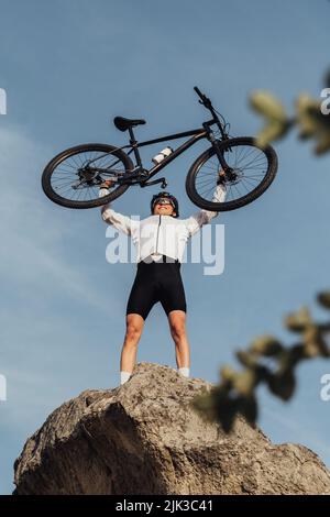 Professioneller Radsportler, der die Hände mit dem Fahrrad hebt, während er auf dem Gipfel des Hügels steht, Himmel auf dem Hintergrund Stockfoto