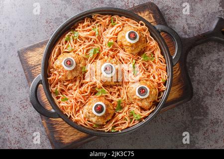 Gruseliges Halloween-Essen aus Fleischbällchen mit Monsteraugen und Pasta mit Tomatensauce in einer Pfanne auf dem Tisch aus der Nähe. Horizontale Draufsicht von oben Stockfoto