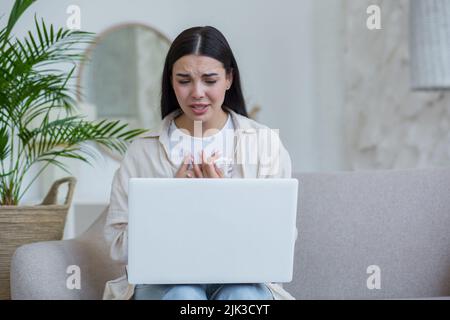 Junge schöne Frau allein zu Hause in Depressionen weinend, Brünette zu Hause sitzen und mit Laptop für Online-Beratungsgespräch mit Psychotherapeuten Stockfoto