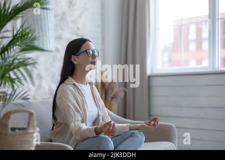Eine schöne Brünette sitzt zu Hause auf der Couch, ruht sich aus, meditiert in der Lotusposition, macht Yoga Stockfoto