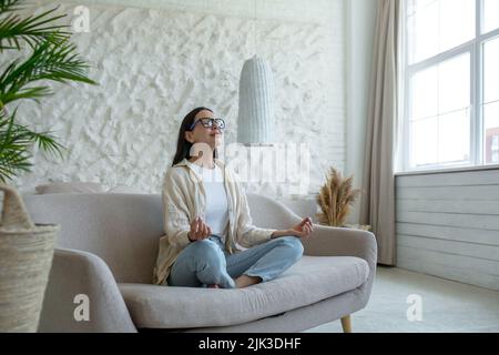 Eine schöne Brünette sitzt zu Hause auf der Couch, ruht sich aus, meditiert in der Lotusposition, macht Yoga Stockfoto