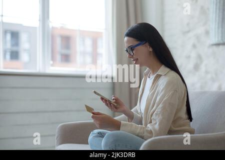 Junge schöne Frau in Brille lächelnd und glücklich beim Online-Shopping im Online-Shop sitzen auf dem Sofa zu Hause, halten Handy und Bankkreditkarte Stockfoto