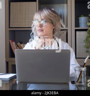 Ärztin vor einem Laptop, der vom Heimstudio aus arbeitet. Moderne Arztpraxis mit Frau in Krankenhausuniform mit Computer auf dem Schreibtisch. Menschen i Stockfoto