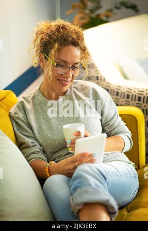 Frau zu Hause viel Spaß und genießen Sie das Lesen von E-Book von modernen Leser. Weibchen haben Freizeitaktivitäten in der Wohnung flach auf einer gelben Couch und Stockfoto