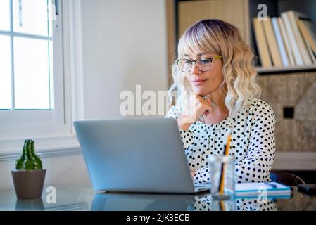 Moderne freiberufliche Unternehmerin Frau, die an einem Laptop im Studio zu Hause arbeitet. Glückliche Frau lächelt vor dem Computer und macht einen Videoanruf Stockfoto
