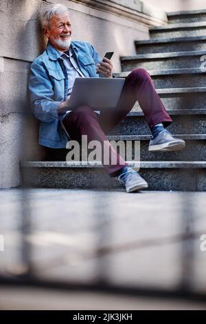 Ein leises Senior Businessman sitzt auf der Treppe draußen und nutzt einen Laptop für die Arbeit. Stockfoto