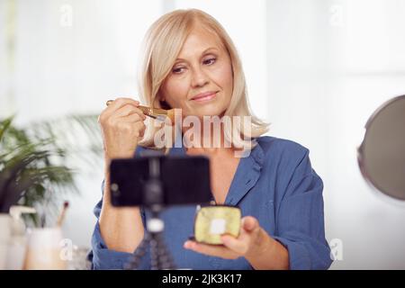 Eine ältere Frau sitzt zu Hause, blush auf und vloggt darüber. Stockfoto