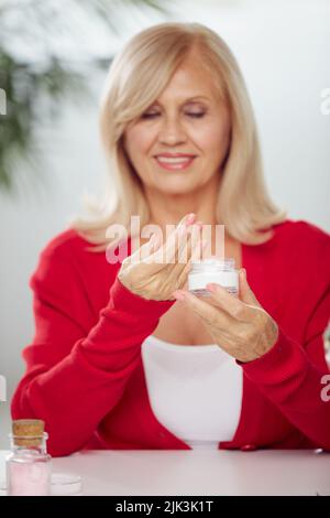 Eine glückliche ältere Frau, die zu Hause sitzt und Anti-Age-Creme auf ihr Gesicht legt. Senioren mit Make-up Stockfoto