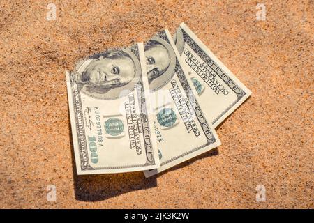 Gelddolare, die halb mit Sand bedeckt sind, liegen in der Nähe des Strandes. Dollarscheine teilweise im Sand vergraben. Dreihundert Dollar im Sand am Meer Ozean Strand begraben Konzept finanzieren Geld Urlaub entspannen Urlaub Stockfoto