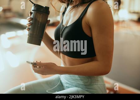 Eine starke, muskulöse Gewichtheber-Frau sitzt in einer Pause im Fitnessstudio, trinkt Proteinshake und hängt am Smartphone. Stockfoto