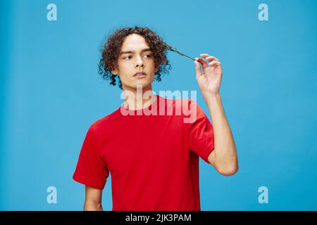 Nachdenklich nachdenklich unglücklich junge lockig gebräunt kaukasischen Kerl in grundlegenden roten T-Shirt posiert isoliert auf blauem Hintergrund. Lifestyle und Emotionen Stockfoto