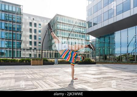 Fröhlicher und gutaussehender Erwachsener, der ein farbenfrohes T-Shirt trägt und akrobatische Trick-Moves in der Stadt macht, alternatives Konzept für Werbung mit energischem, p Stockfoto