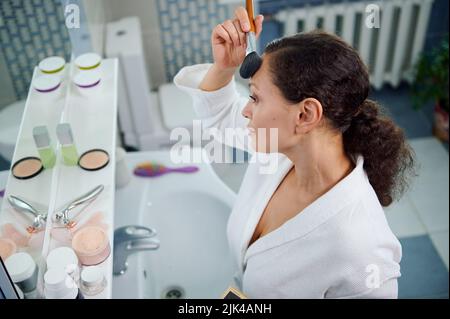 Draufsicht. Entzückende Frau, die ein mattierendes Pulver auf ihr Gesicht aufsetzt, zufrieden mit ihrem Aussehen und ihrem natürlichen Make-up Stockfoto