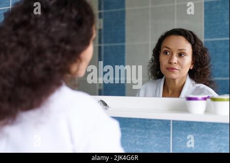 Schöne lockige Brünette, hispanische Frau in weißem Bademantel, reflektiert im Spiegel, während sie sich das Badezimmer bewundert Stockfoto